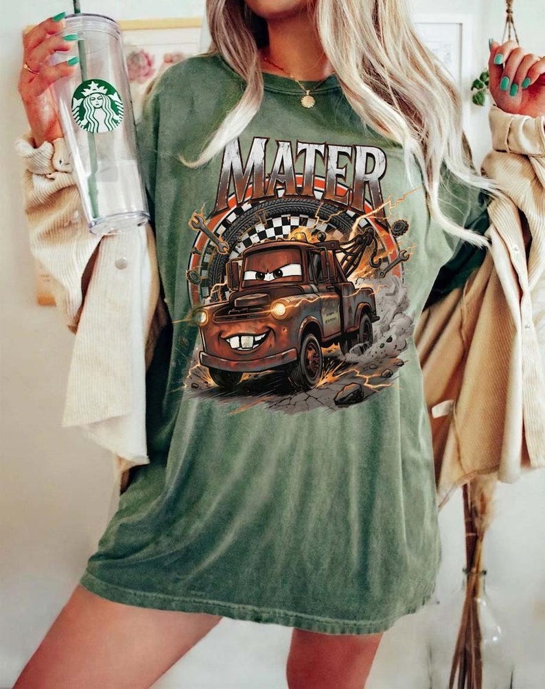 Pu&ograve; includere: T-shirt verde con la scritta "MATER" e un'illustrazione del personaggio Mater del film Cars. Il design include un carro attrezzi, una bandiera a scacchi e degli attrezzi. La maglietta ha un aspetto vintage e sbiadito, che le conferisce un aspetto usurato.