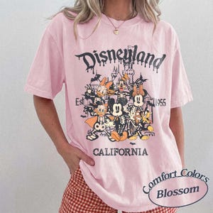 May include: Pink t-shirt with a Disneyland graphic featuring cartoon characters in skeleton costumes. The design includes the text "Disneyland California Est 1955" in a gothic font. The shirt is a Comfort Colors Blossom.