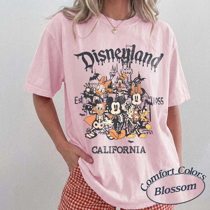 May include: Pink t-shirt with a Disneyland graphic featuring cartoon characters in skeleton costumes. The design includes the text "Disneyland California Est 1955" in a gothic font. The shirt is a Comfort Colors Blossom.