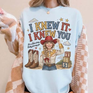 May include: A light blue crewneck sweatshirt featuring a graphic print of a person in a cowboy hat, boots, and western shirt. The text reads I KNEW IT, I KNEW YOU, It's a Love story Baby just say yes, and T S 5 on stacked blocks with stars.