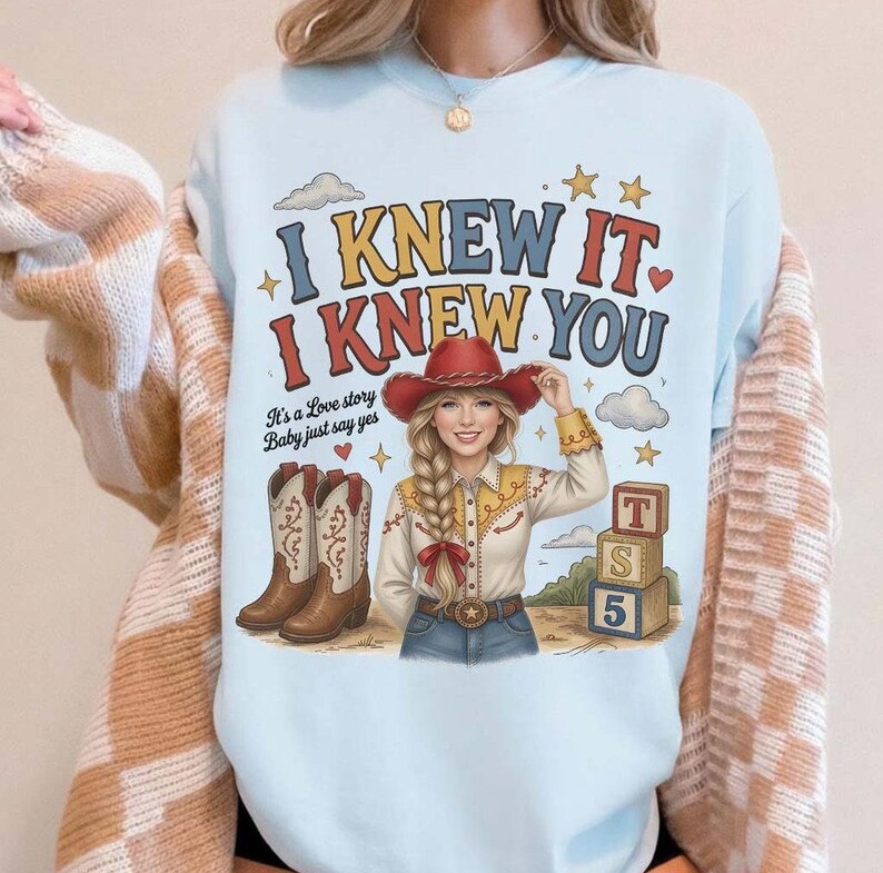 May include: A light blue crewneck sweatshirt featuring a graphic print of a person in a cowboy hat, boots, and western shirt. The text reads I KNEW IT, I KNEW YOU, It's a Love story Baby just say yes, and T S 5 on stacked blocks with stars.