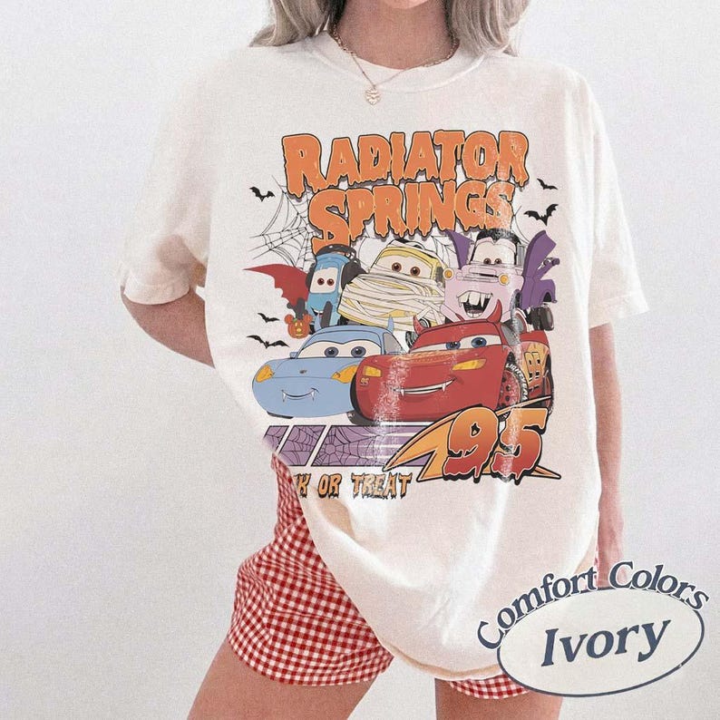 May include: Ivory-colored t-shirt featuring a Halloween-themed graphic of characters from the movie Cars, with the text "Radiator Springs" and "95". The design includes bats, a spider web, and the phrase "Trick or Treat".
