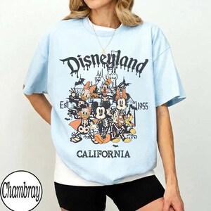 May include: Light blue t-shirt with a Disneyland graphic. The graphic features Disney characters in skeleton costumes, a castle, and the text "Disneyland California Est. 1955".