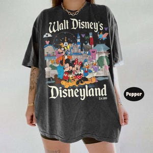 Puede incluir: Camiseta gris oscuro con un gr&aacute;fico de Disneyland con Mickey Mouse, Minnie Mouse, Donald Duck y otros personajes de Disney. La camiseta tiene el texto "Walt Disney's Disneyland" y "Est. 1955".