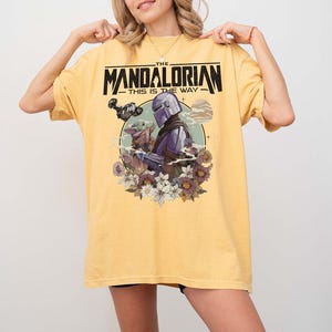 May include: Yellow t-shirt featuring a graphic from The Mandalorian. The design includes the text "THE MANDALORIAN - THIS IS THE WAY" above an illustration of the Mandalorian and Grogu surrounded by flowers. The shirt has short sleeves.