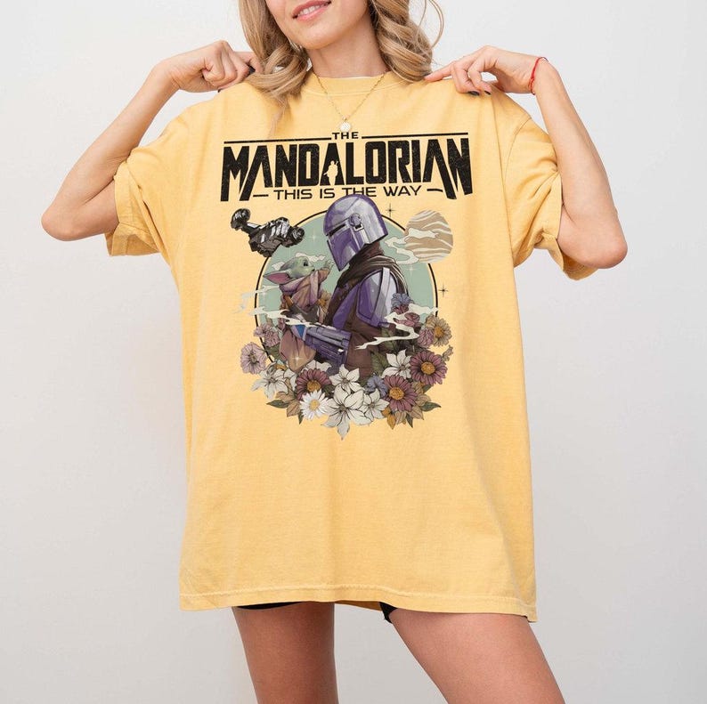 May include: Yellow t-shirt featuring a graphic from The Mandalorian. The design includes the text "THE MANDALORIAN - THIS IS THE WAY" above an illustration of the Mandalorian and Grogu surrounded by flowers. The shirt has short sleeves.