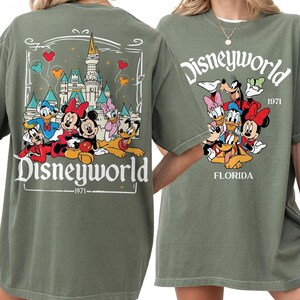 May include: Olive green t-shirt featuring a Disney World graphic. The front design includes the word "Disneyworld" above a group of cartoon characters and the text "1971 FLORIDA". The back design features a castle and Disney characters.