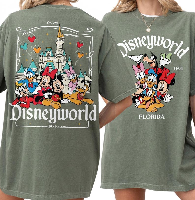 May include: Olive green t-shirt featuring a Disney World graphic. The front design includes the word "Disneyworld" above a group of cartoon characters and the text "1971 FLORIDA". The back design features a castle and Disney characters.