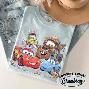 Pode incluir: Camiseta Chambray Comfort Colors azul claro com carros de desenho animado com orelhas de coelhinho da P&aacute;scoa e decora&ccedil;&otilde;es florais. O design inclui Rel&acirc;mpago McQueen, Mate e outros personagens, al&eacute;m de ovos e flores de P&aacute;scoa.