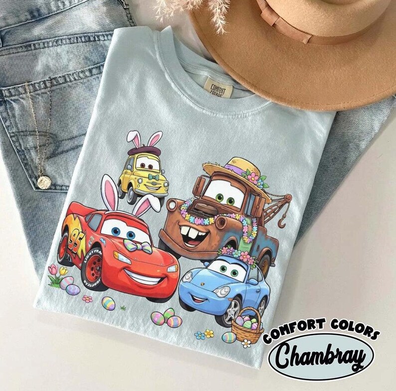 Pode incluir: Camiseta Chambray Comfort Colors azul claro com carros de desenho animado com orelhas de coelhinho da P&aacute;scoa e decora&ccedil;&otilde;es florais. O design inclui Rel&acirc;mpago McQueen, Mate e outros personagens, al&eacute;m de ovos e flores de P&aacute;scoa.