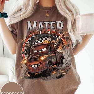 Pu&ograve; includere: T-shirt color espresso con grafica di Mater del film Cars. Il design include la scritta "MATER" sopra il personaggio, con una bandiera a scacchi e attrezzi sullo sfondo. La maglietta ha un aspetto invecchiato e vintage.