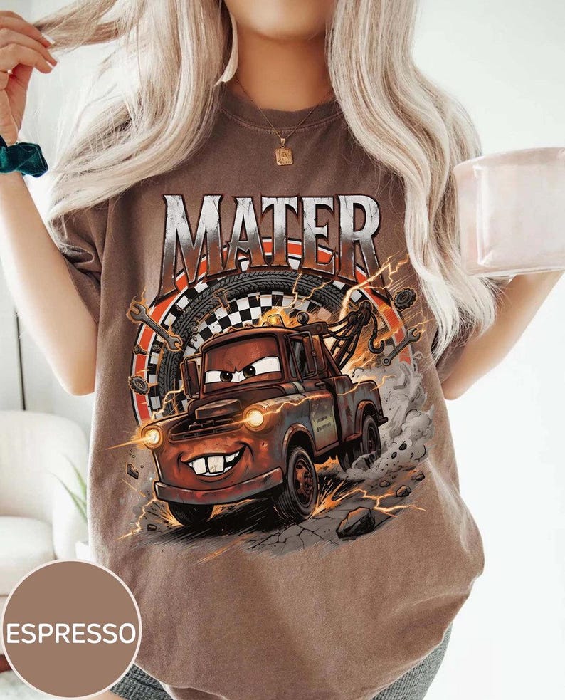 Pu&ograve; includere: T-shirt color espresso con grafica di Mater del film Cars. Il design include la scritta "MATER" sopra il personaggio, con una bandiera a scacchi e attrezzi sullo sfondo. La maglietta ha un aspetto invecchiato e vintage.