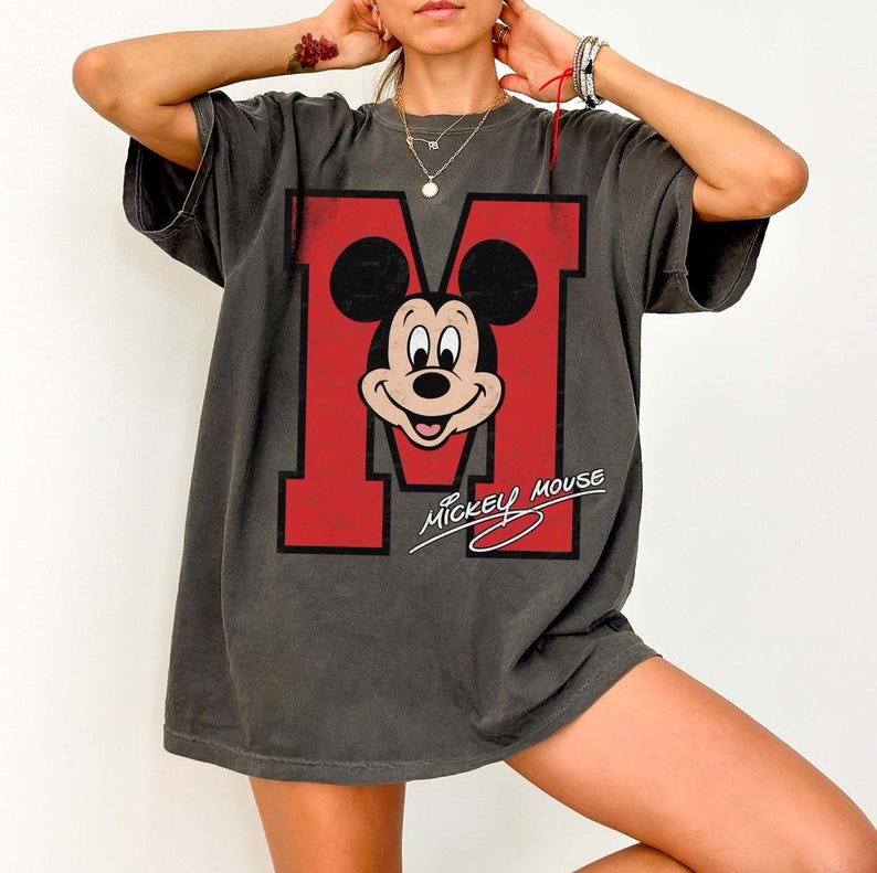 May include: Dark gray t-shirt featuring a large red letter "M" with Mickey Mouse's face in the center. The words "Mickey Mouse" are written in a cursive font below the graphic. The shirt has short sleeves and a relaxed fit.