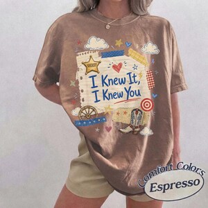 Op de afbeelding: Een bruin t-shirt met een grafische print van een vel gelinieerd papier met de tekst "I Knew It, I Knew You" in het blauw. Het ontwerp bevat een gouden sheriffster, een cowboylaars, een wagenwiel, een rood hart, wolken en sterren in diverse kleuren.