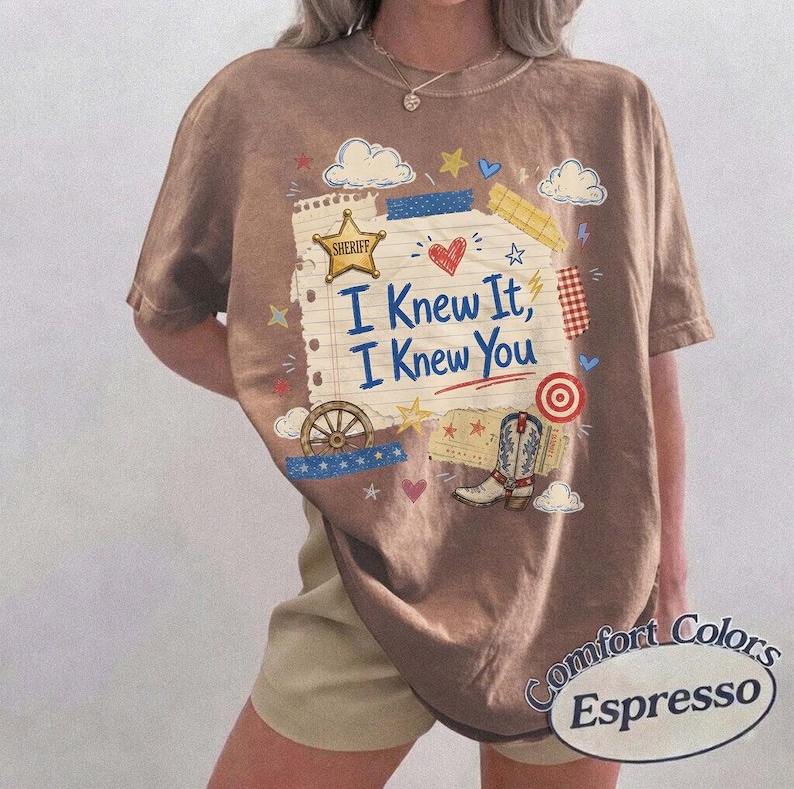 Op de afbeelding: Een bruin t-shirt met een grafische print van een vel gelinieerd papier met de tekst "I Knew It, I Knew You" in het blauw. Het ontwerp bevat een gouden sheriffster, een cowboylaars, een wagenwiel, een rood hart, wolken en sterren in diverse kleuren.