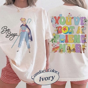 May include: An ivory t-shirt featuring a graphic of Bo Peep with her staff and the text Bo Peep on the front. The back displays colorful cartoon characters from Toy Story with the text You've Got A Friend In Me in large, multi-colored bubble letters.