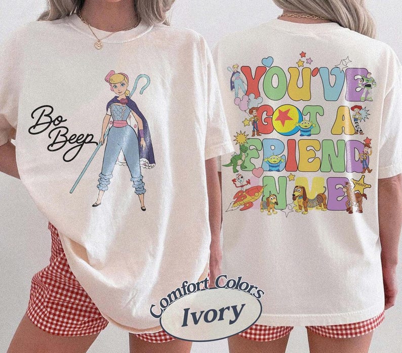 May include: An ivory t-shirt featuring a graphic of Bo Peep with her staff and the text Bo Peep on the front. The back displays colorful cartoon characters from Toy Story with the text You've Got A Friend In Me in large, multi-colored bubble letters.