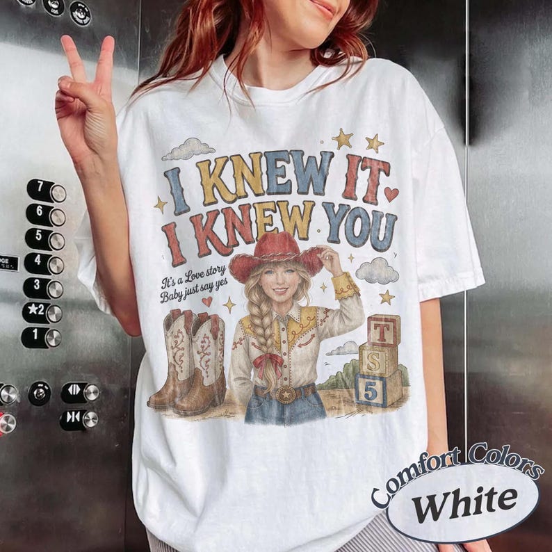 May include: A white t-shirt featuring a graphic print with the text I KNEW IT I KNEW YOU and It's a Love story Baby just say yes. The design includes a person in a cowboy hat, a pair of western boots, stars, clouds, and three stacked blocks.