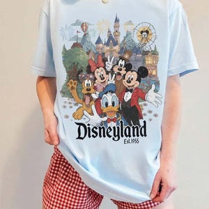 Pu&ograve; includere: Una t-shirt azzurro chiaro con una stampa grafica di Topolino, Minnie, Paperino, Pippo e Pluto davanti al castello di Disneyland. Il testo "Disneyland Est. 1955" &egrave; stampato in nero sotto i personaggi. La t-shirt &egrave; abbinata a pantaloncini a quadretti vichy rossi e bianchi.