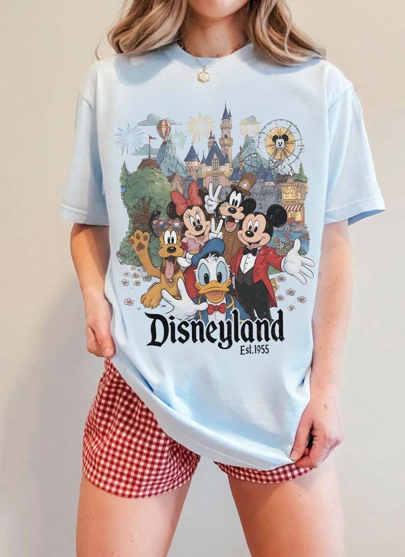 Pu&ograve; includere: Una t-shirt azzurro chiaro con una stampa grafica di Topolino, Minnie, Paperino, Pippo e Pluto davanti al castello di Disneyland. Il testo "Disneyland Est. 1955" &egrave; stampato in nero sotto i personaggi. La t-shirt &egrave; abbinata a pantaloncini a quadretti vichy rossi e bianchi.