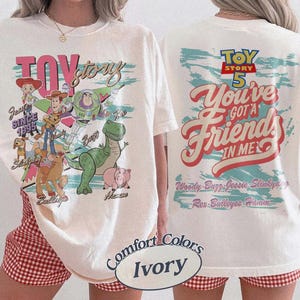 Pode incluir: Uma t-shirt cor de marfim com gr&aacute;ficos das personagens de Toy Story. A frente mostra a Jessie, o Woody, o Buzz Lightyear, o Slinky Dog, o Bullseye, o Rex e o Hamm com o texto Toy Story. As costas apresentam o log&oacute;tipo Toy Story 5 e o texto You've Got A Friend In Me.