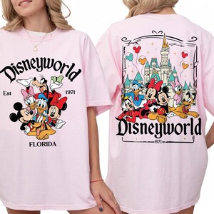 May include: Pink t-shirt featuring a Disneyworld design. The front displays Mickey Mouse, Minnie Mouse, Donald Duck, Daisy Duck, Goofy, and Pluto. The back shows the Disney castle and characters. Text includes "Disneyworld", "Est 1971", and "Florida".