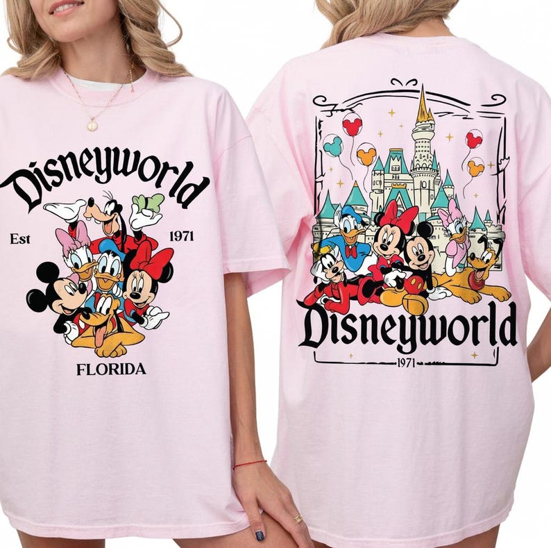 May include: Pink t-shirt featuring a Disneyworld design. The front displays Mickey Mouse, Minnie Mouse, Donald Duck, Daisy Duck, Goofy, and Pluto. The back shows the Disney castle and characters. Text includes "Disneyworld", "Est 1971", and "Florida".