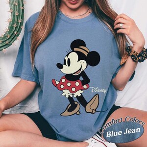May include: Blue jean Comfort Colors t-shirt featuring a classic Minnie Mouse graphic. Minnie is depicted in black, white, and red, with a polka-dot skirt and tan shoes. The Disney logo is visible.