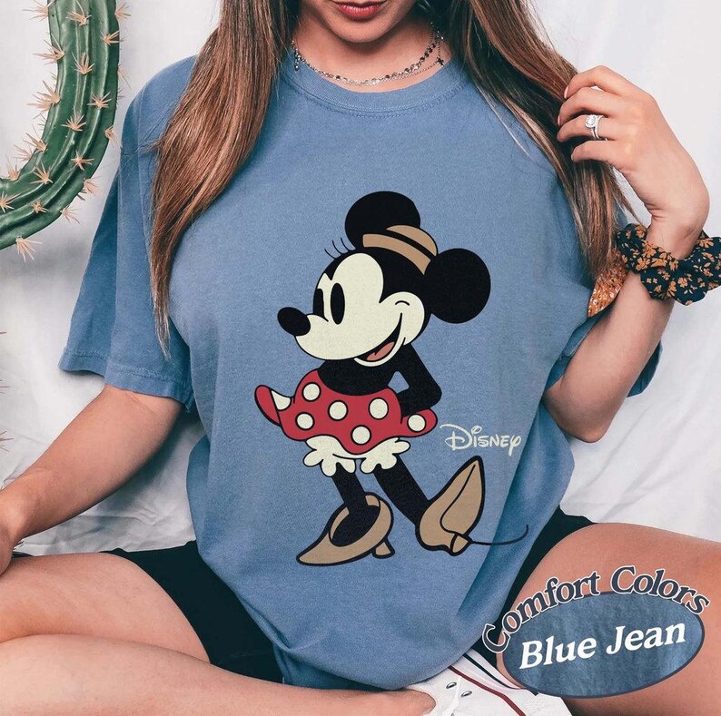 May include: Blue jean Comfort Colors t-shirt featuring a classic Minnie Mouse graphic. Minnie is depicted in black, white, and red, with a polka-dot skirt and tan shoes. The Disney logo is visible.