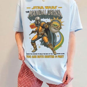 May include: A light blue t-shirt featuring a graphic of The Mandalorian and Grogu. The text reads STAR WARS THE MANDALORIAN, BOUNTY HUNTER, and WHEN YOU CHOOSE TO WALK THE WAY OF THE MANDALORE YOU ARE BOTH HUNTER & PREY in yellow and black lettering.