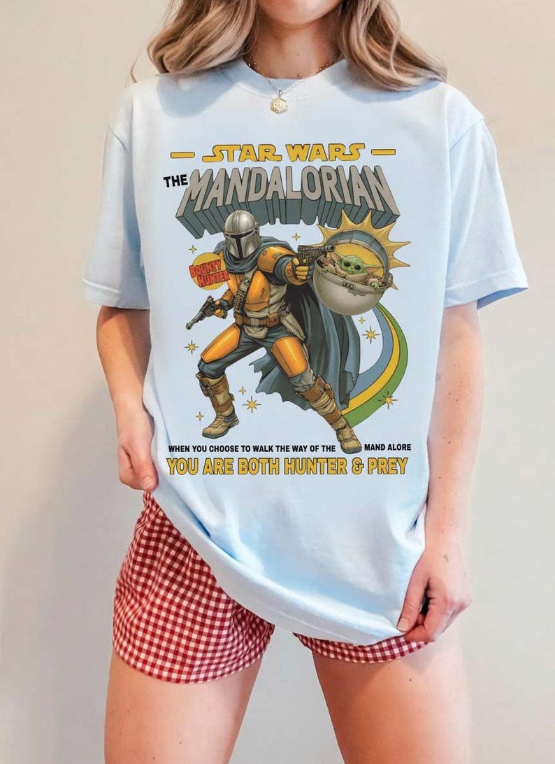 May include: A light blue t-shirt featuring a graphic of The Mandalorian and Grogu. The text reads STAR WARS THE MANDALORIAN, BOUNTY HUNTER, and WHEN YOU CHOOSE TO WALK THE WAY OF THE MANDALORE YOU ARE BOTH HUNTER & PREY in yellow and black lettering.