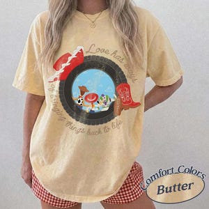 Może przedstawiać: Bladoż&oacute;łty t-shirt z kr&oacute;tkim rękawem z grafiką czarnej opony otaczającej scenę z niebieskim niebem i postaciami z kresk&oacute;wek. Tekst brzmi Love has ways of bringing things back to life. Widoczny jest r&oacute;wnież czerwony kowbojski kapelusz i czerwony but.