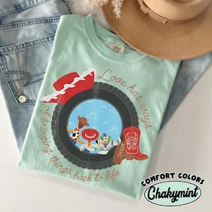 Może przedstawiać: Jasnozielony t-shirt w kolorze miętowym z grafiką czarnej opony otaczającej scenę z niebieskim niebem i postaciami z kresk&oacute;wek. Wz&oacute;r zawiera czerwony kowbojski kapelusz, czerwony but oraz napis Love has ways of bringing things back to life.