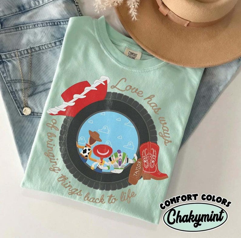 Może przedstawiać: Jasnozielony t-shirt w kolorze miętowym z grafiką czarnej opony otaczającej scenę z niebieskim niebem i postaciami z kresk&oacute;wek. Wz&oacute;r zawiera czerwony kowbojski kapelusz, czerwony but oraz napis Love has ways of bringing things back to life.