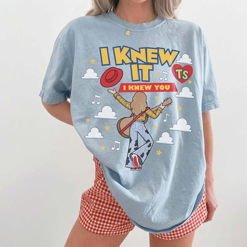 May include: A light blue t-shirt featuring a graphic of a person playing an acoustic guitar, surrounded by clouds, stars, and musical notes. The text reads I KNEW IT and I KNEW YOU, accompanied by a red hat, a red heart with TS, and cow print pants.