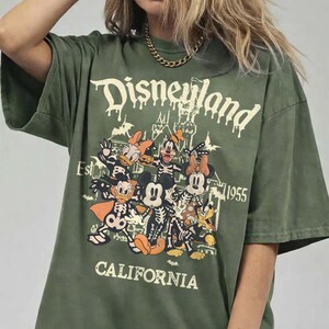 May include: Olive green t-shirt with a Disneyland graphic featuring cartoon characters in skeleton costumes. The text "Disneyland California Est. 1955" is printed in white. The shirt has short sleeves and a relaxed fit.