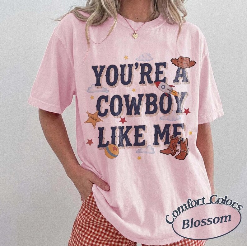以下が含まれることがあります： YOU'RE A COWBOY LIKE MEという濃い青色の文字がプリントされたライトピンクの半袖Tシャツ。茶色のカウボーイハット、ロケット、星、ボール、茶色のカウボーイブーツのイラストが描かれており、小さな星がアクセントになっています。