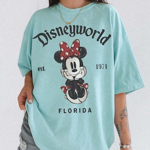May include: A light blue t-shirt featuring a graphic of Minnie Mouse with a red polka dot bow. The shirt has the text "Disneyworld" above the graphic, with "est. 1971" and "FLORIDA" below.