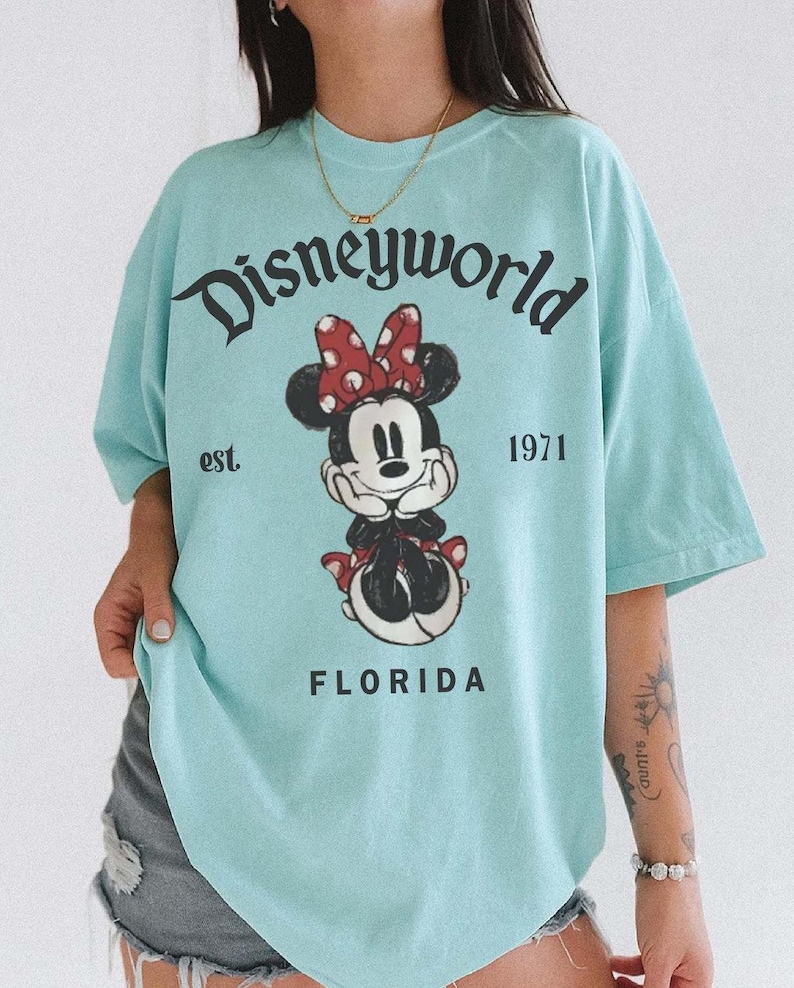 May include: A light blue t-shirt featuring a graphic of Minnie Mouse with a red polka dot bow. The shirt has the text "Disneyworld" above the graphic, with "est. 1971" and "FLORIDA" below.