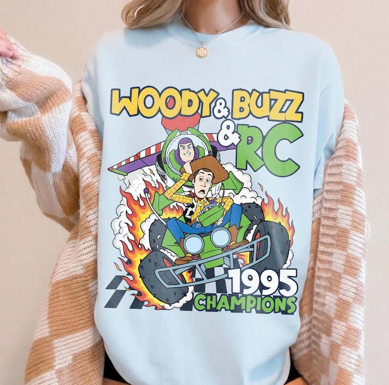May include: Light blue sweatshirt featuring a graphic of Woody, Buzz Lightyear, and RC from Toy Story. The design includes the text "WOODY & BUZZ & RC 1995 CHAMPIONS" in yellow and green. The image shows Woody in a car engulfed in flames.