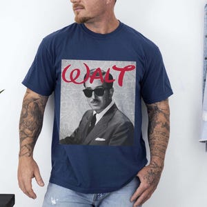 May include: Navy blue t-shirt featuring a black and white image of a man wearing sunglasses, with the name "WALT" in red script above the image. The shirt is a classic crew neck style.