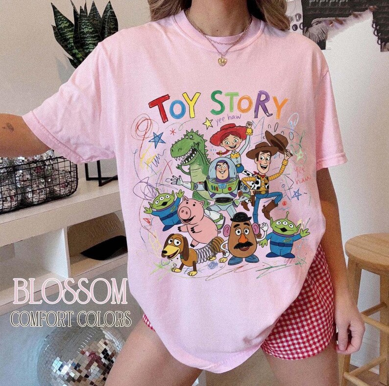 May include: A light pink t-shirt featuring a colorful graphic of characters from the animated film "Toy Story." The design includes the words "Toy Story" in a playful font, along with the phrase "yee haw." The shirt also has the text "Blossom Comfort Colors."