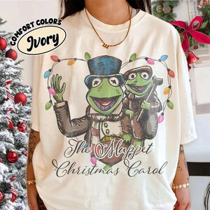 May include: An ivory t-shirt featuring a graphic of two green frog characters dressed in Victorian-style clothing, surrounded by a string of colorful Christmas lights. The text "The Muppet Christmas Carol" is printed below in a black script font.