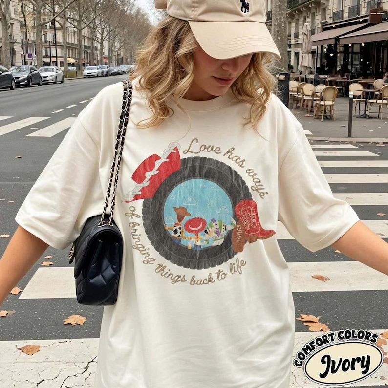 Może przedstawiać: Kremowy t-shirt z kr&oacute;tkim rękawem z grafiką przedstawiającą oponę z niebieskim środkiem, czerwony kowbojski kapelusz i czerwony kowbojski but. Tekst brzmi, Love has ways of bringing things back to life. Widoczna jest czarna pikowana torebka na ramię.