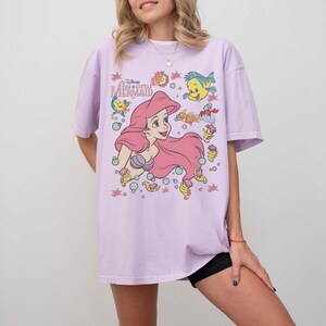 May include: A light purple t-shirt featuring a graphic of Ariel from Disney's The Little Mermaid. The design includes Ariel with pink hair, Flounder, and other sea creatures. The text "The Little Mermaid" is also visible.