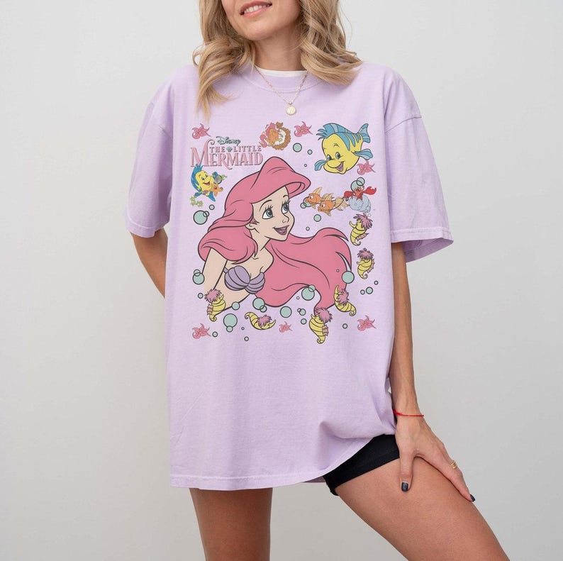 May include: A light purple t-shirt featuring a graphic of Ariel from Disney's The Little Mermaid. The design includes Ariel with pink hair, Flounder, and other sea creatures. The text "The Little Mermaid" is also visible.