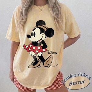 May include: Yellow t-shirt featuring a Minnie Mouse graphic. The cartoon character is depicted in black, white, and red, with the Disney logo. The shirt is labeled "Comfort Colors Butter".