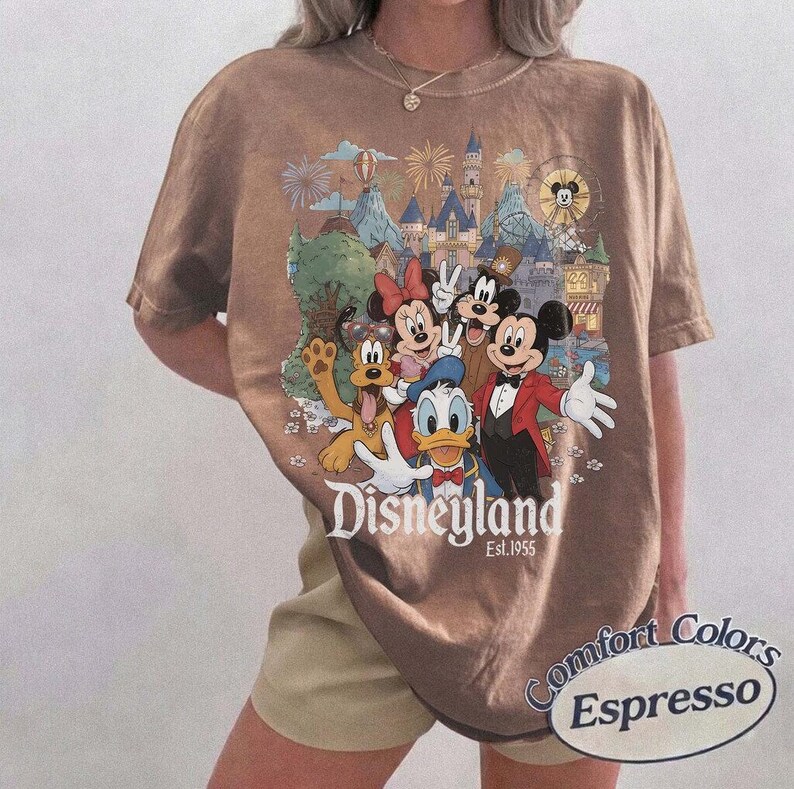 Pu&ograve; includere: Una t-shirt marrone a maniche corte con una stampa grafica di Topolino, Minni, Paperino, Pippo e Pluto davanti a un castello con fuochi d'artificio. Il testo "Disneyland Est. 1955" &egrave; stampato sotto i personaggi. La maglietta &egrave; etichettata Comfort Colors Espresso.