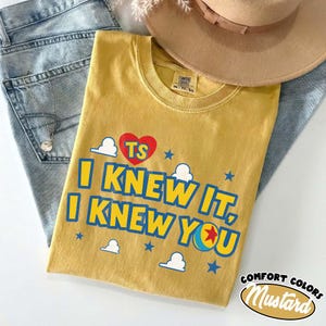 Puede incluir: Una camiseta de cuello redondo color amarillo mostaza con un coraz&oacute;n rojo con las letras TS, texto azul que dice I KNEW IT, I KNEW YOU, y un gr&aacute;fico de una pelota con una estrella roja, rodeado de nubes blancas y estrellas azules peque&ntilde;as.