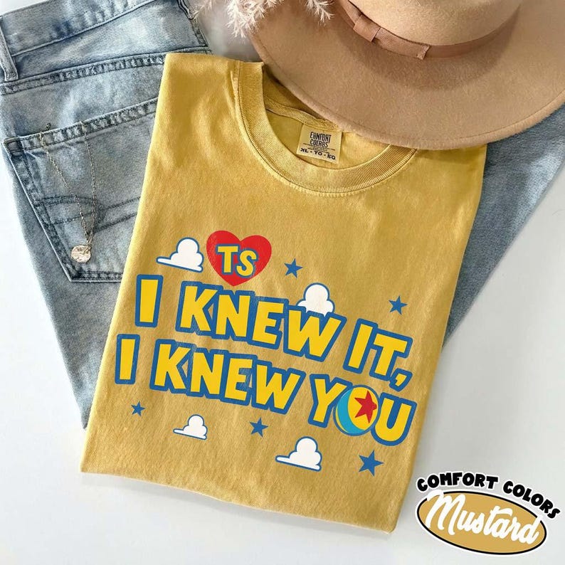Puede incluir: Una camiseta de cuello redondo color amarillo mostaza con un coraz&oacute;n rojo con las letras TS, texto azul que dice I KNEW IT, I KNEW YOU, y un gr&aacute;fico de una pelota con una estrella roja, rodeado de nubes blancas y estrellas azules peque&ntilde;as.